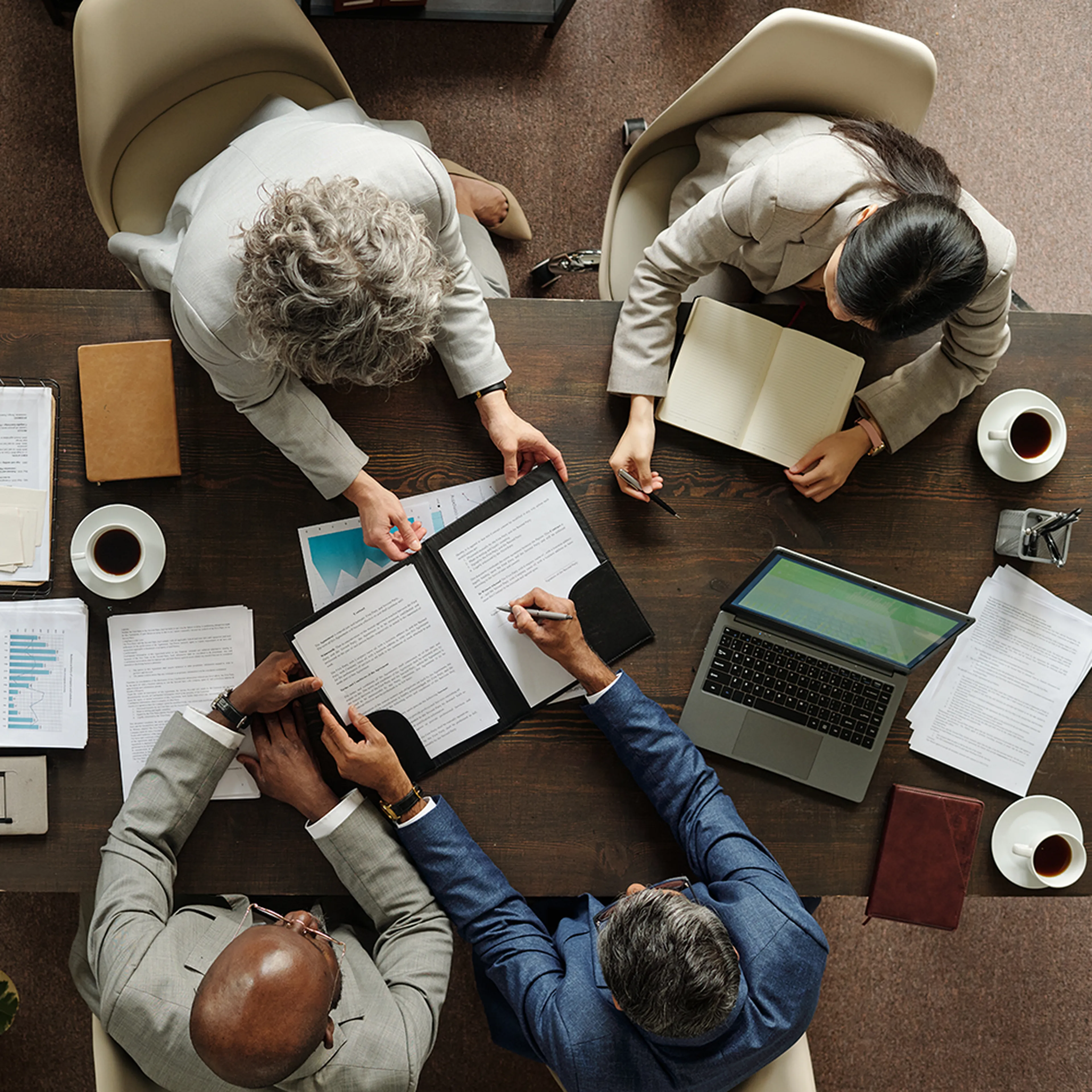 Four people in a meeting around a table with documents, a laptop, notebooks, and coffee cups. They are discussing and reviewing papers.