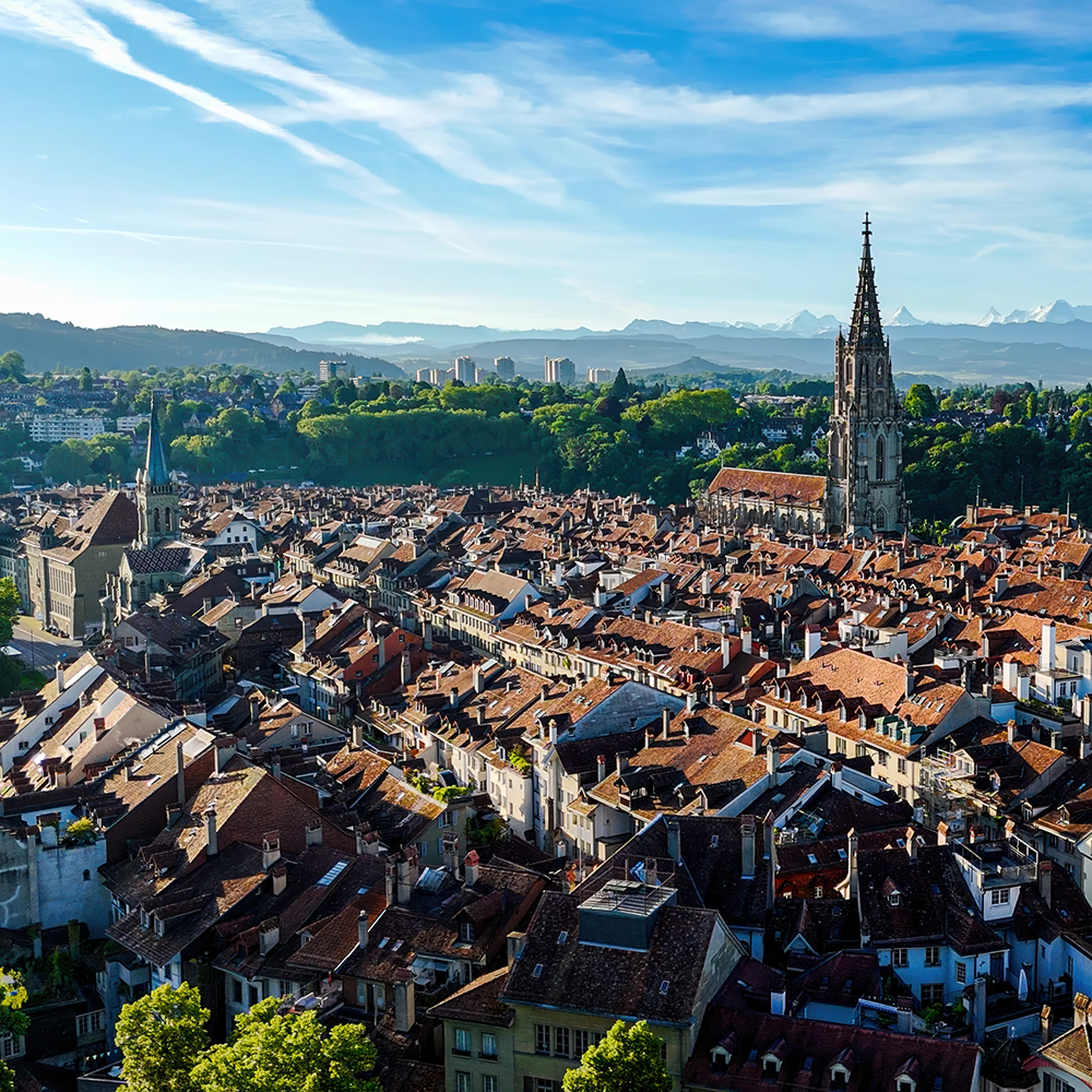 Aerial view of Bern, Switzerland, featuring red-roofed buildings, the Bern Cathedral, lush greenery, and distant mountains under a blue sky.