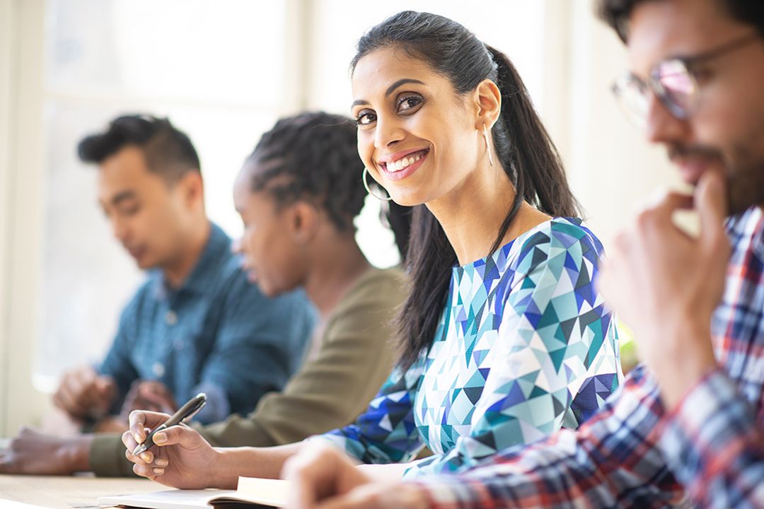 Smiling woman in a blue patterned top sits at a table during a group meeting or class, with three people blurred in the background.
