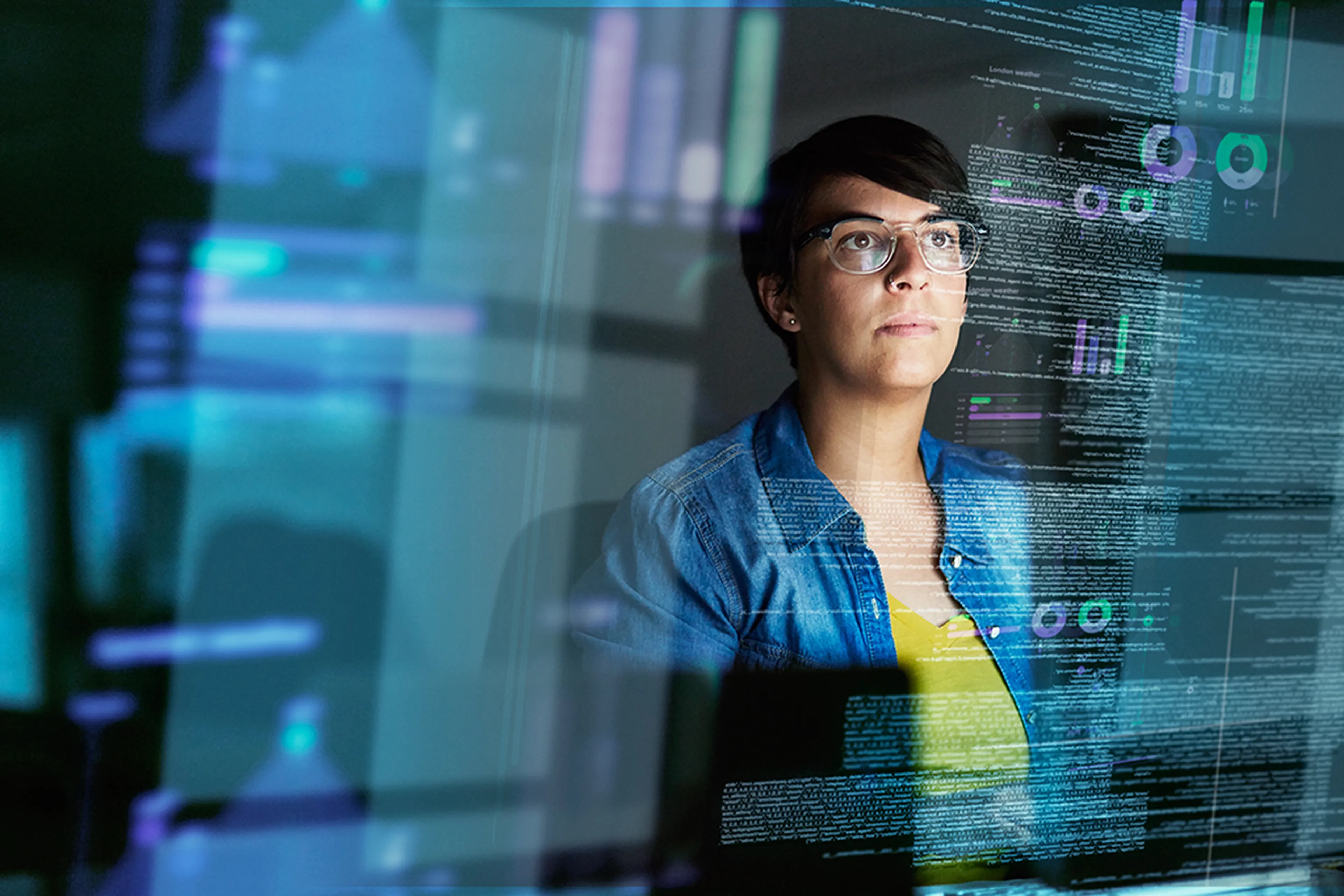 A focused person wearing glasses looks at multiple transparent digital screens displaying data and graphs.