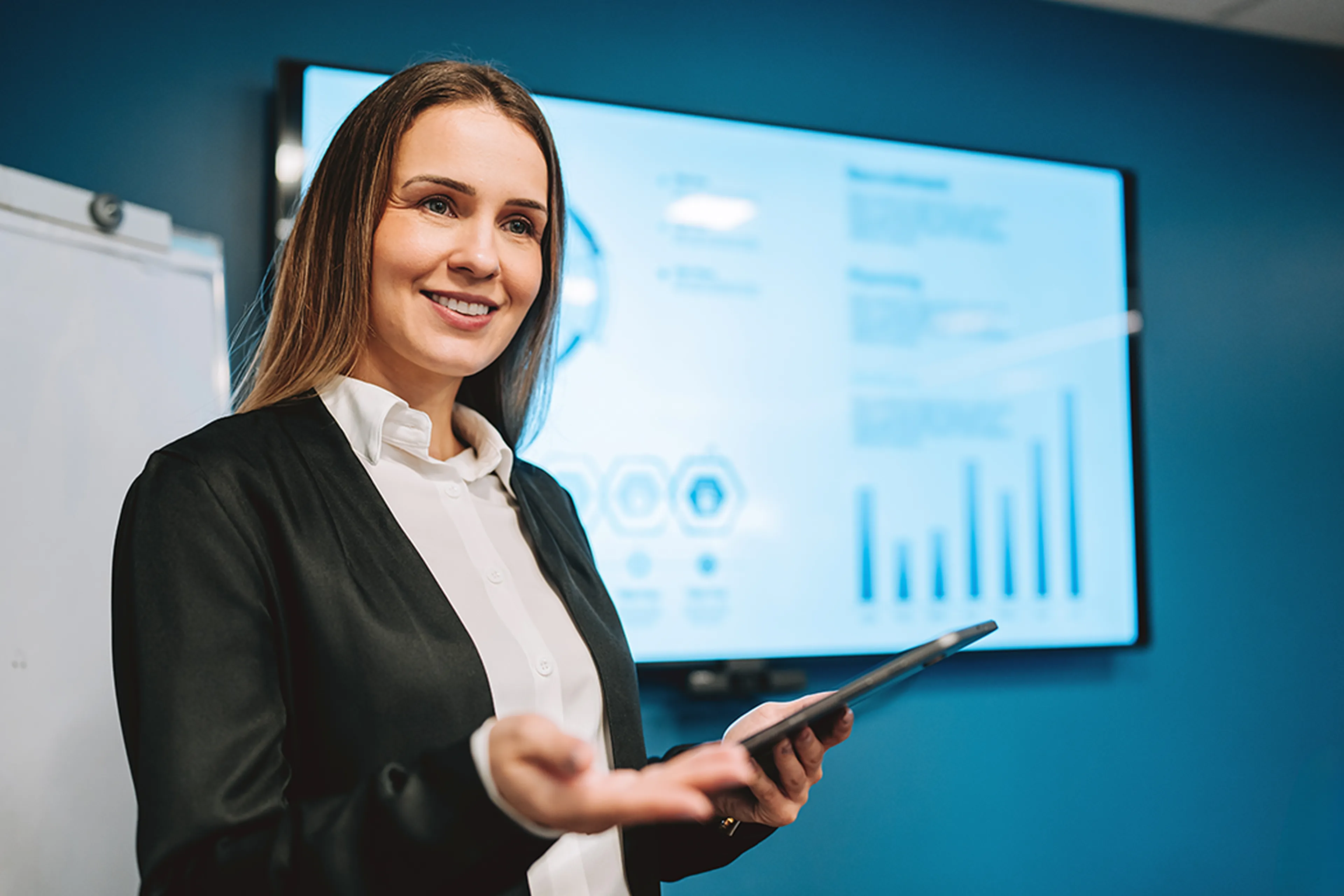 Smiling woman presenting with a tablet in hand, standing in front of a screen displaying graphs and charts in a modern office setting.