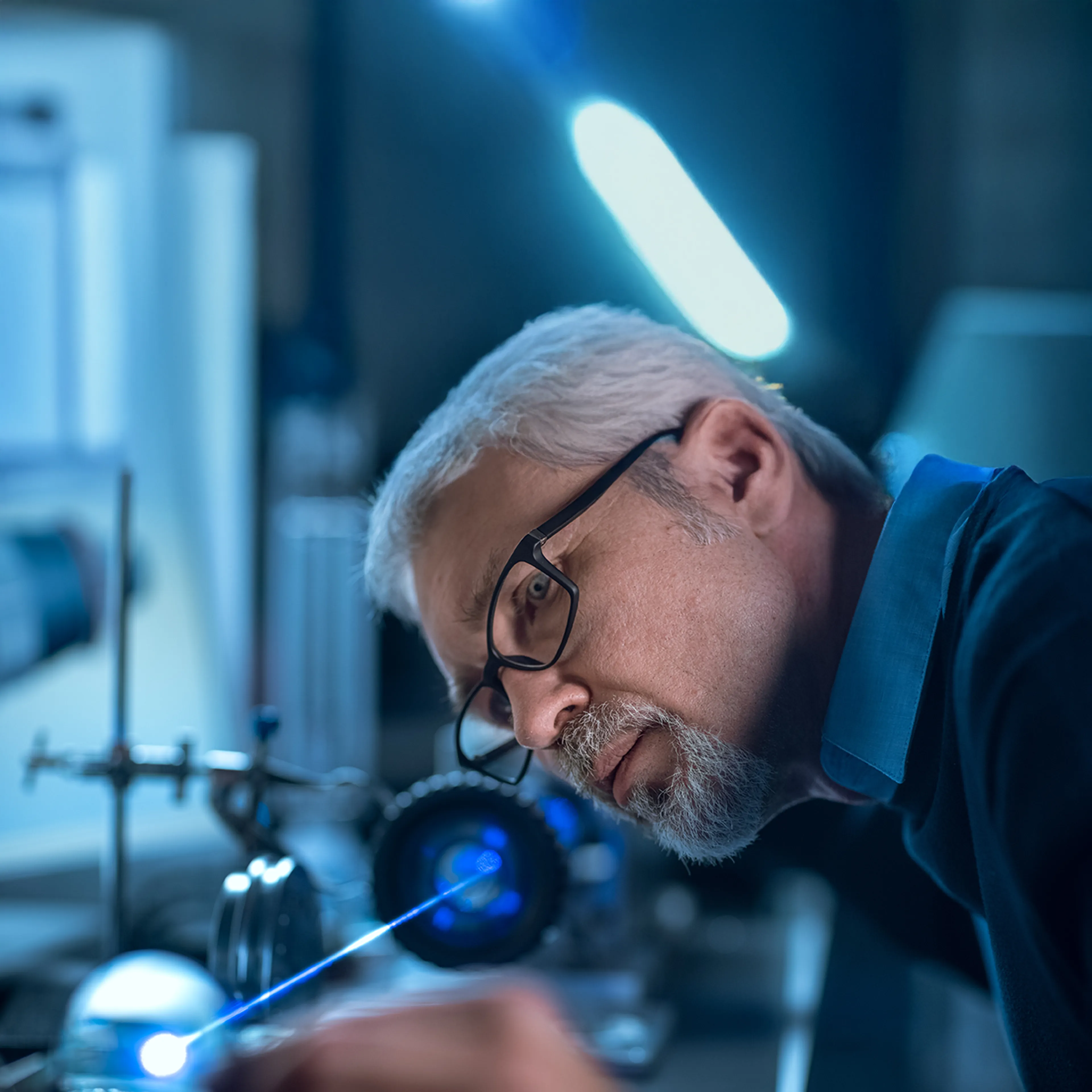 Man with glasses working intently in a dimly lit lab, adjusting equipment with a visible blue laser beam.