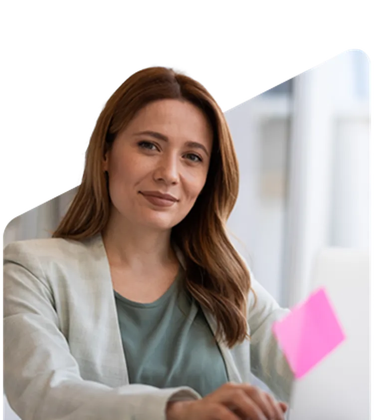 A woman with long hair, wearing a light blazer, sits at a desk with a laptop. A pink sticky note is visible on the screen.