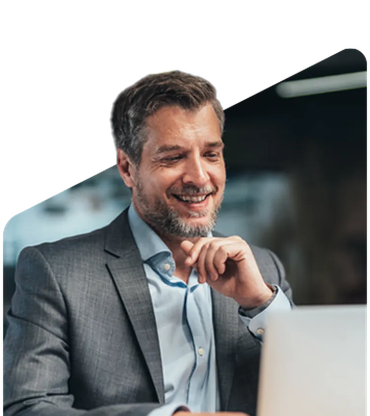 Smiling man in a suit working on a laptop in a dimly lit room, with a hand resting on his chin.