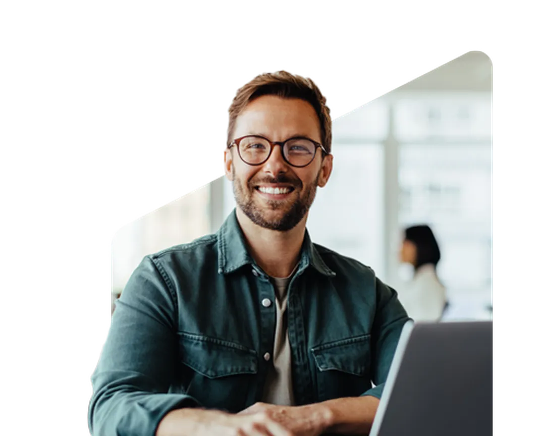 Smiling man with glasses using a laptop, sitting in a bright room with blurred person in the background.