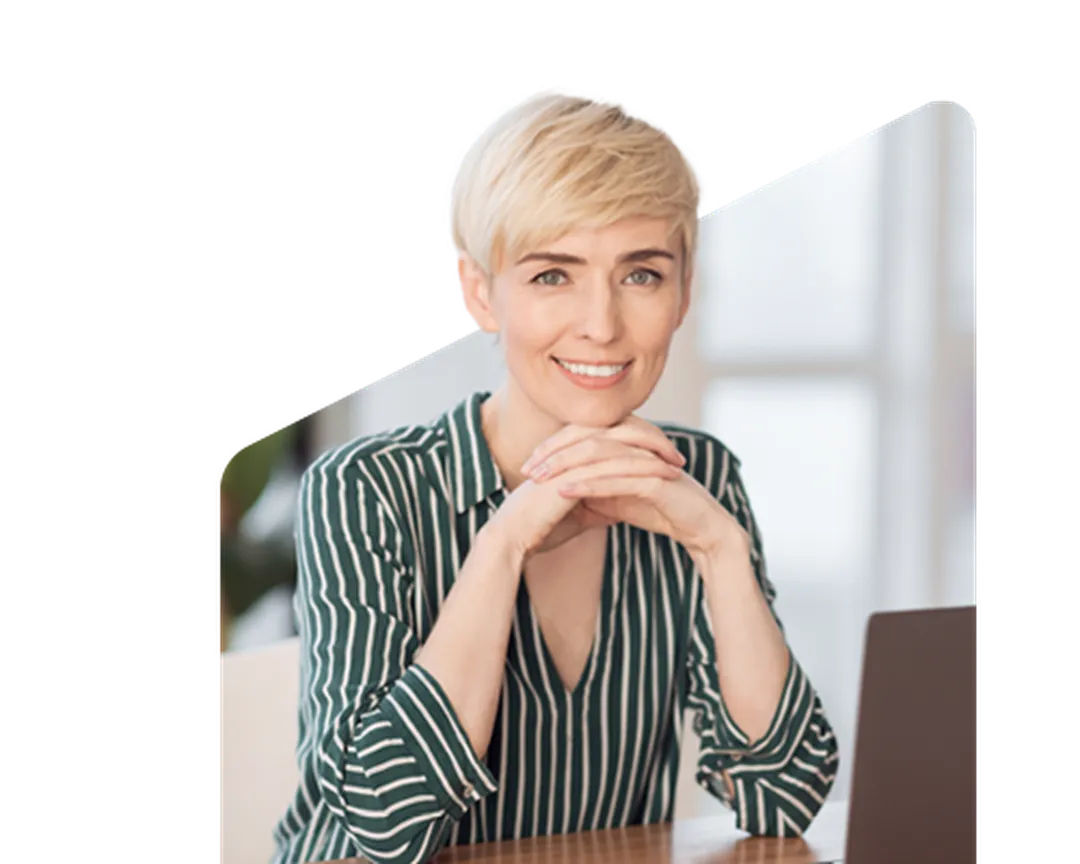 Person with short blonde hair and striped shirt, smiling while sitting at a desk with a laptop in a bright room.