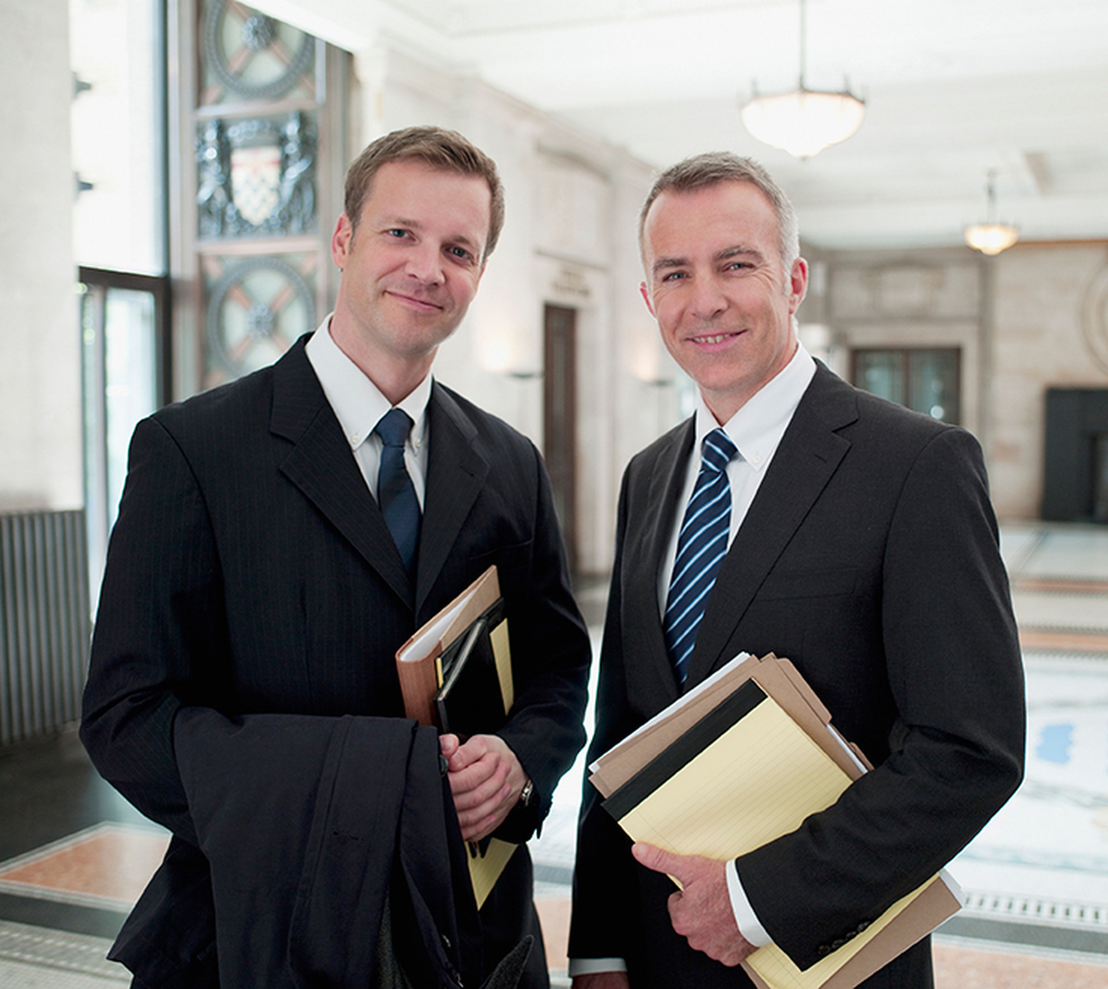 Two lawyers in suits smiling, holding folders and a coat, standing in a bright, spacious hallway with decorative windows and chandeliers.