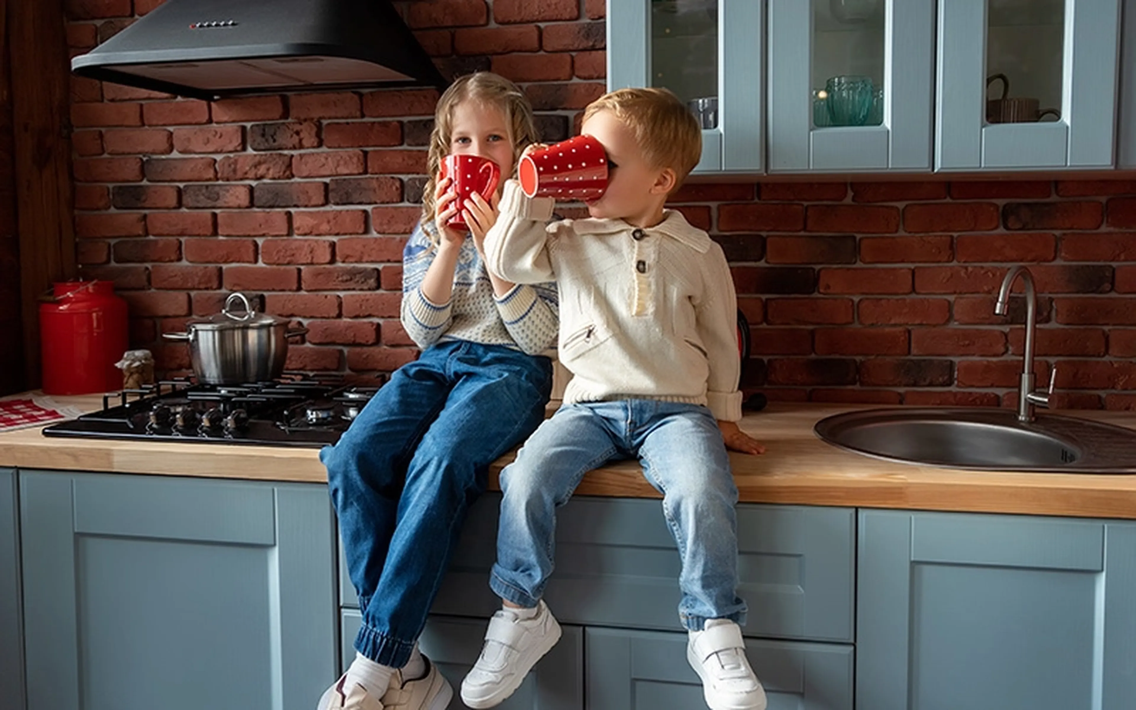 A young brother and sister have fun sitting on a kitchen countertop, drinking from oversized mugs.