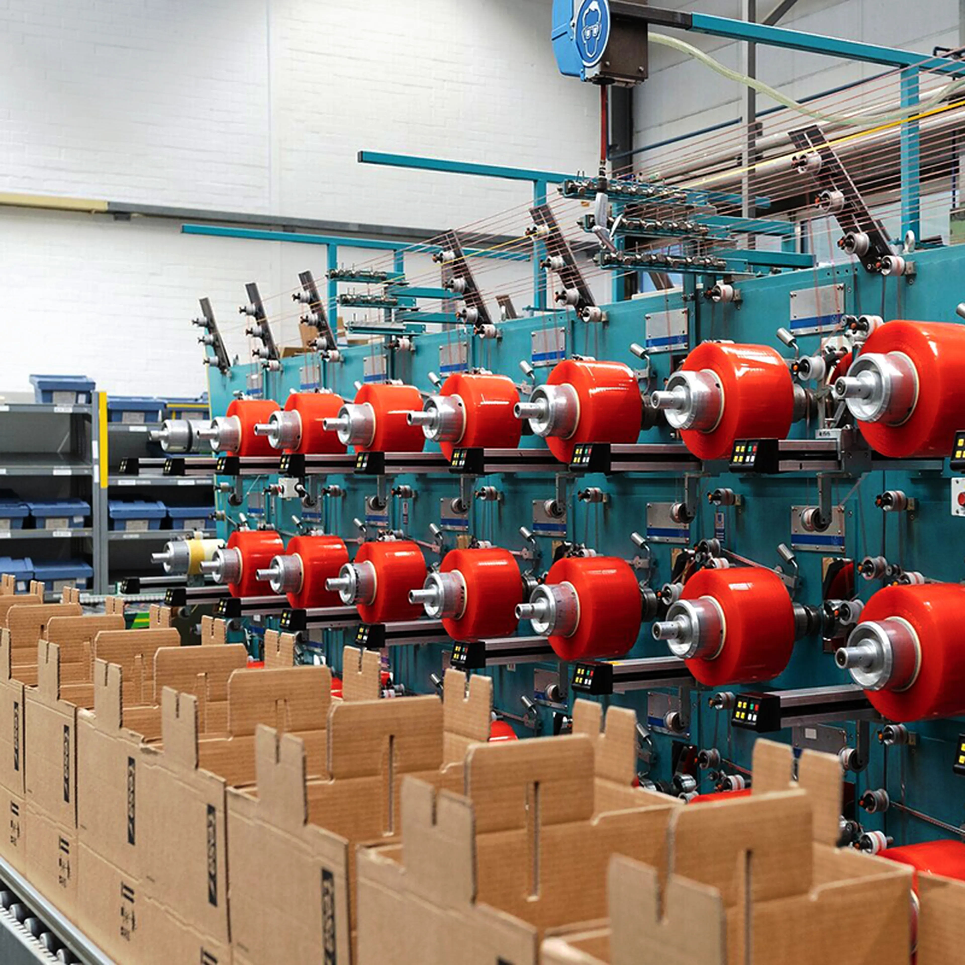 A row of industrial textile machines with red spools in a factory setting, with cardboard boxes in the foreground.