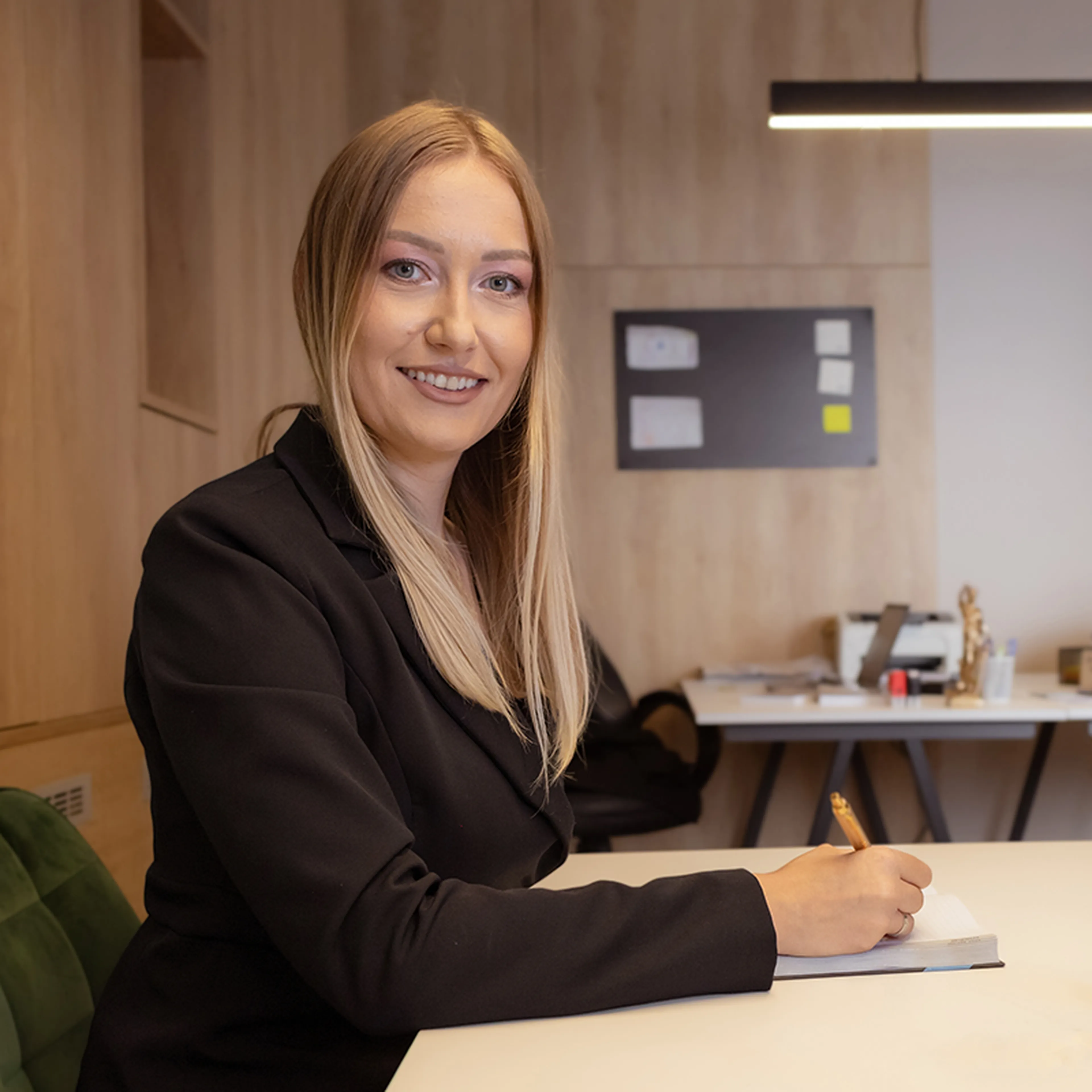 Smiling woman with long blonde hair in a black blazer writing in a notebook at a desk in a modern office setting.