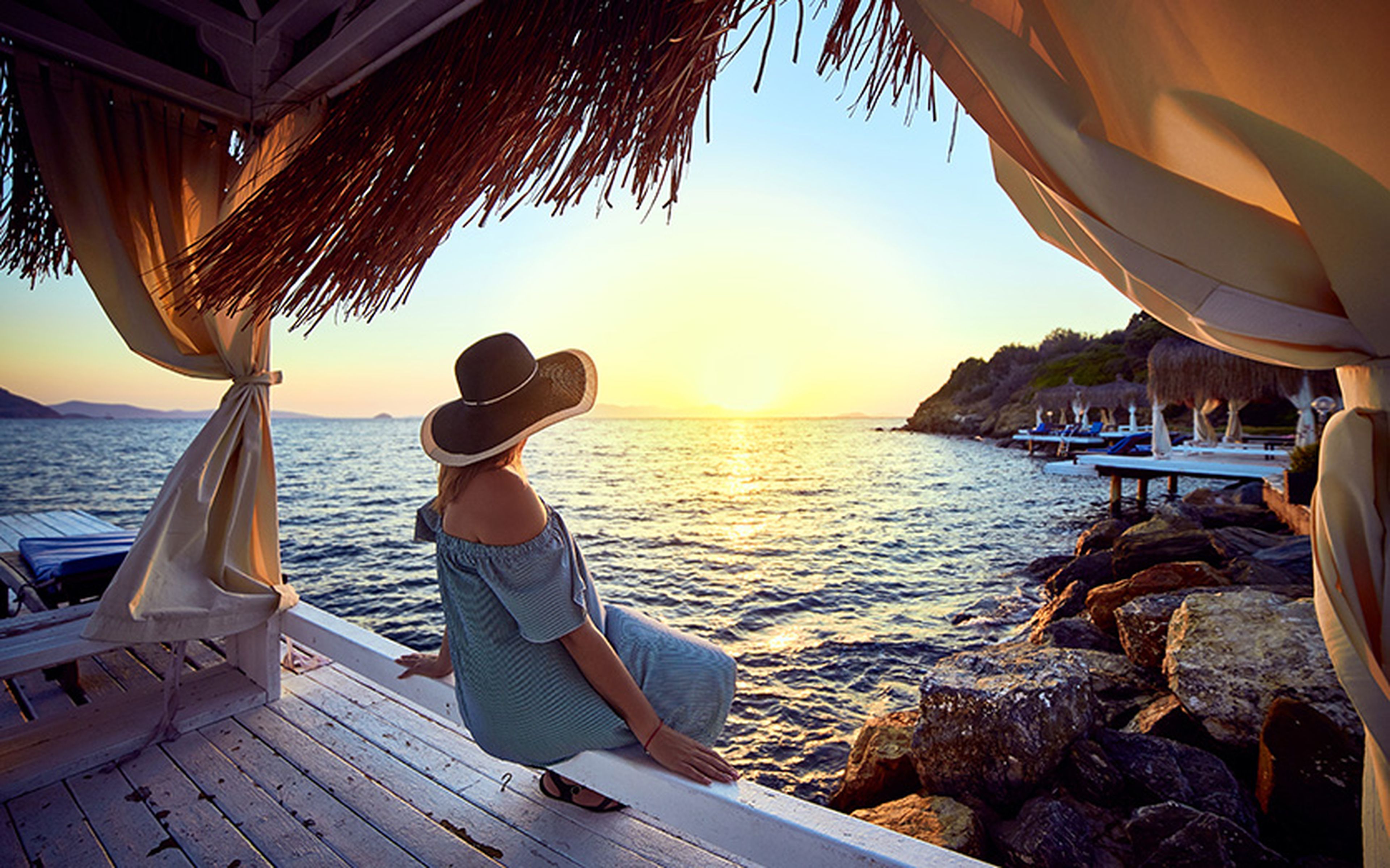 Woman in a wide-brimmed hat sitting by the ocean at a luxury beachfront resort, watching the sunset.