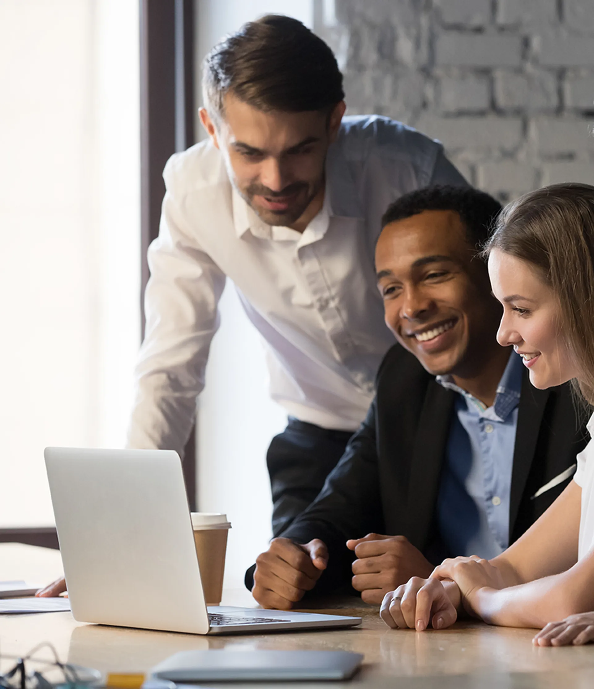Young team smiling and looking at a laptop in an office setting. A coffee cup is on the table next to them.