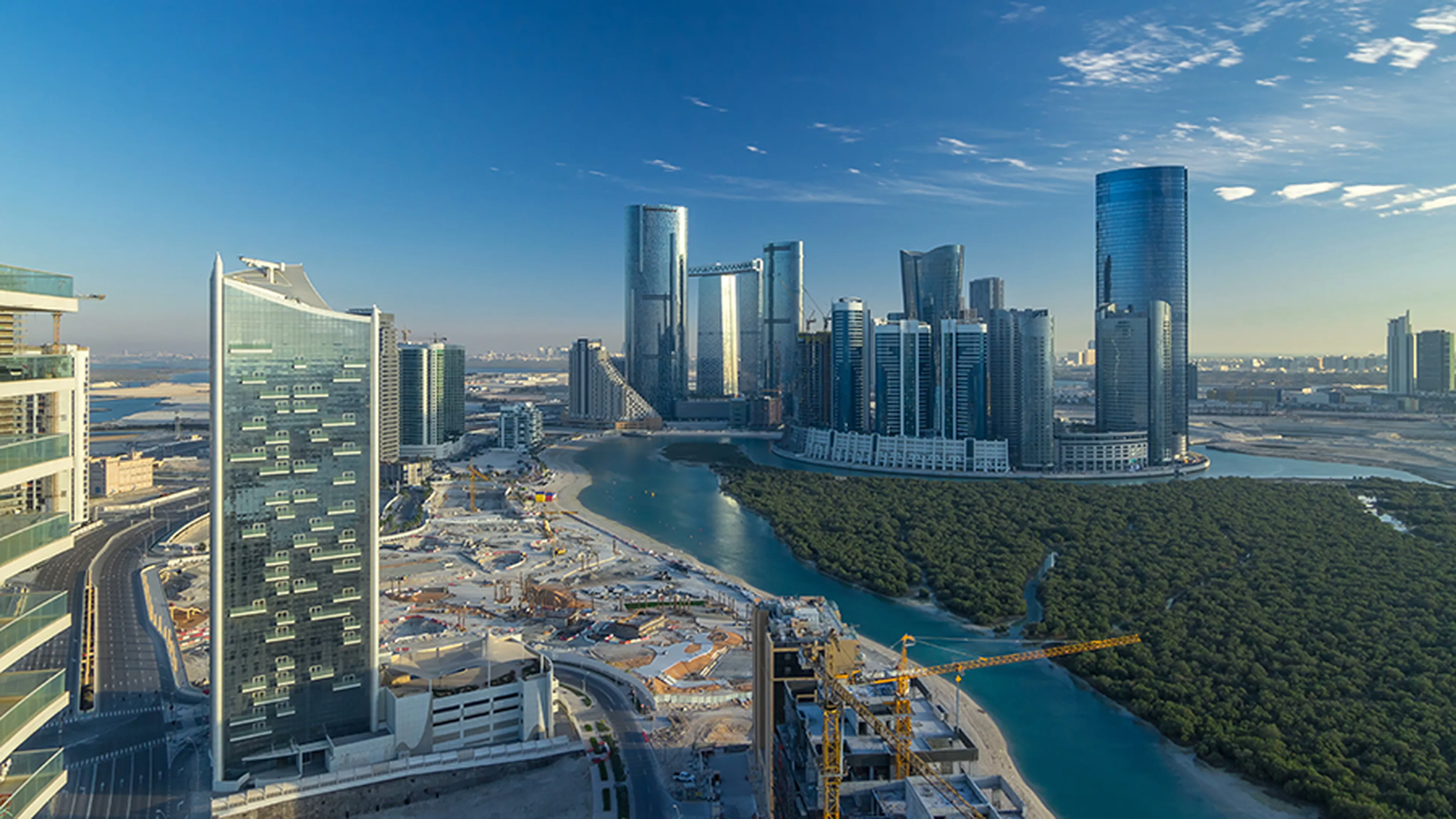 Skyline of Abu Dhabi featuring modern skyscrapers, a winding river, construction area, and lush greenery under a clear blue sky.