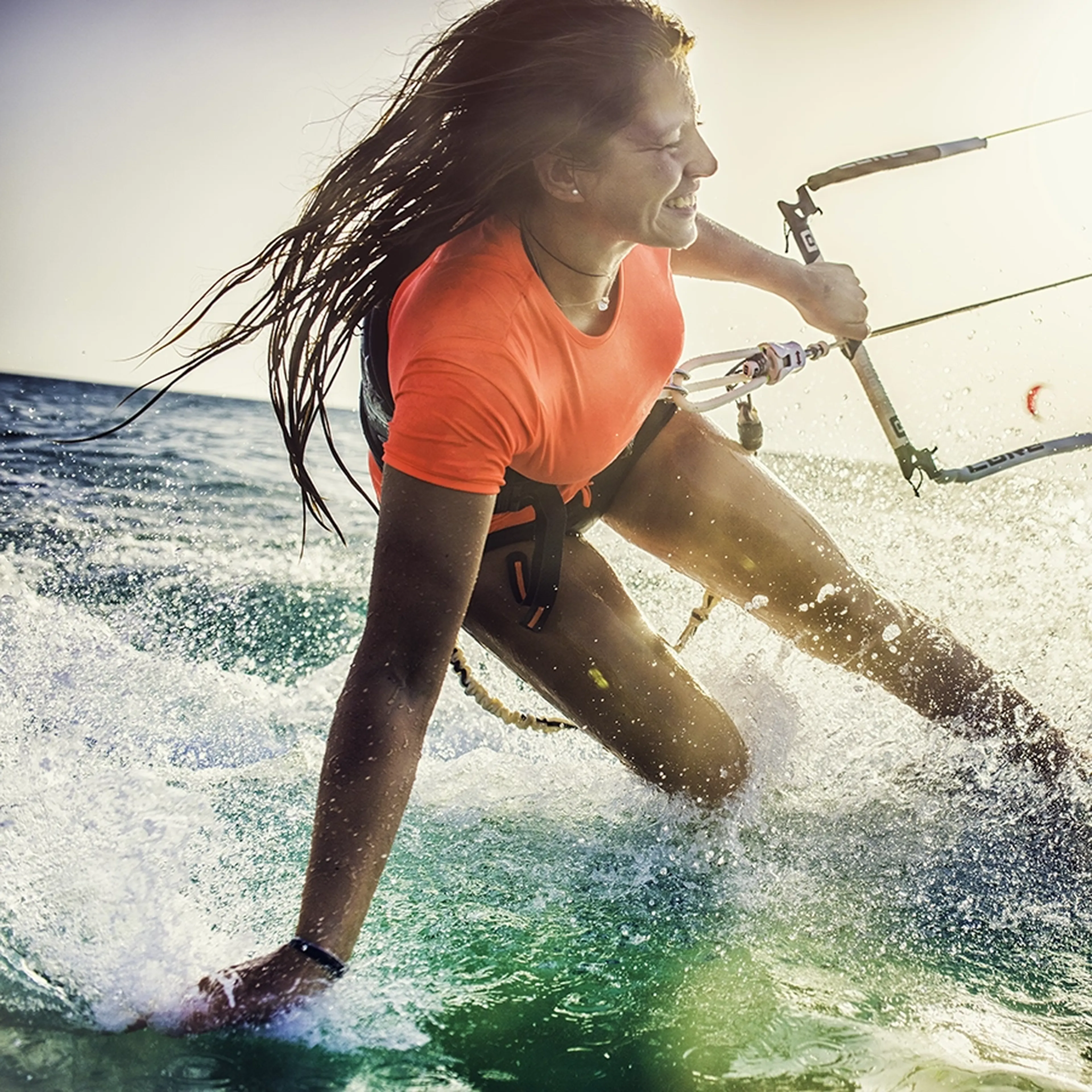Woman in a bright shirt kitesurfing, splashing water, with a joyful expression in the sunlight over the ocean.